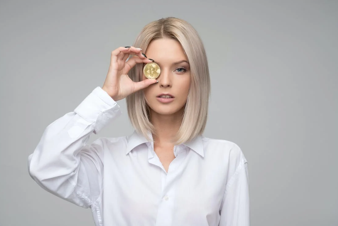 a woman in a white shirt is standing, holding a Bitcoin coin in her eye